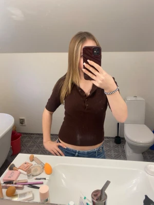 Brun ribbad croppad polotopp - Snygg brun ribbad topp med korta ärmar och knappar framtill. Modellen är croppad och har en klassisk polokrage. Perfekt att matcha med jeans för en trendig look.