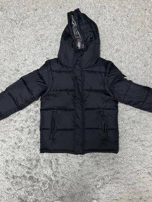 Svart pufferjacka från Burberry - Säljer en svart pufferjacka från Burberry med huva och dragkedja. Jackan har två fickor med dragkedja och Burberry-logga på ärmen. Insidan har klassiskt rutigt Burberry-mönster. Perfekt för kalla vinterdagar och riktigt snygg streetstil.