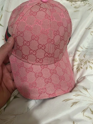 Rosa Gucci keps med monogram - Snygg rosa keps från Gucci med klassiskt GG-monogram över hela kepsen. Kepsen har böjd skärm, knapp på toppen och detaljer i grönt och rött på sidan. Tillverkad i canvasmaterial med exklusiv känsla. Perfekt för dig som vill sticka ut med en trendig accessoar.