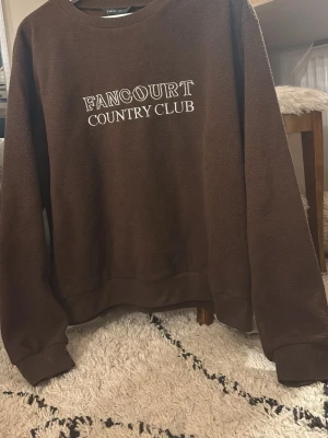 Brun fleece-tröja från SHEIN storlek 38 - Jag är en Xs och den är perfekt om man vill ha en lite oversize tröja. Mysig brun fleece-tröja med vit brodyrtext 'Fancourt Country Club' på bröstet. Tröjan har rund halsringning, långa ärmar och ribbade muddar. Perfekt för en avslappnad och sportig stil. 