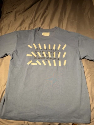 Maison Margiela blå t-shirt med print - Blå t-shirt från Maison Margiela med ikoniskt grafiskt print och logga på bröstet. Tillverkad i mjuk bomull som känns skön mot huden. Perfekt för dig som gillar stilrena men samtidigt unika plagg. Frågor? Tveka inte på att skriva. Mvh Noah OBS, KÖP INTE MED INSTABOX❌