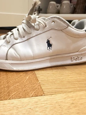 Vita Polo Ralph Lauren sneakers - Klassiska vita sneakers från Polo Ralph Lauren med marinblå logga på sidan och mörkblå detalj på hälen. Skorna har lågt skaft, vita snören och är tillverkade i slätt läder. Perfekta för en clean och stilren look.