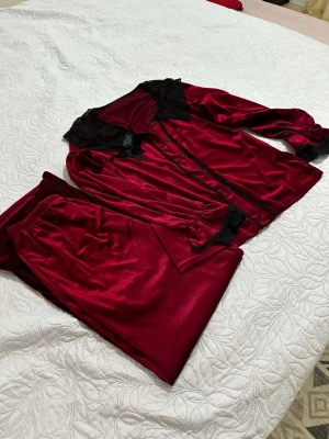 Vinröd sammets pyjamas med spetsdetaljer - Säljer en lyxig vinröd pyjamas i mjuk sammet med svarta spetsdetaljer vid krage och ärmslut. Setet består av en långärmad skjorta med knappar och matchande vida byxor med resår i midjan. Perfekt för mysiga kvällar hemma.
