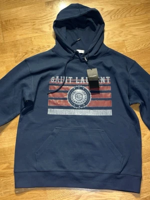 Blå hoodie Saint laur - Säljer en mörkblå hoodie från med tryck i rött, vitt och blått framtill. Hoodien har huva med dragsko och en stor magficka. Tillverkad i mjuk bomull och har en avslappnad passform. Perfekt för dig som gillar exklusiva streetwear-plagg. Passar Även i M