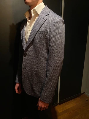 Fiskbensmönstrad Van Gils kavaj - Blå, stickad kavaj från Van Gils i fiskbensmönster med patch pockets. Kavajen är i nyskick, endast använd någon enstaka gång. Stl 48 (stor i storleken, sitter likt Stl 50).                Hör av dig vid eventuella frågor och/eller funderingar!