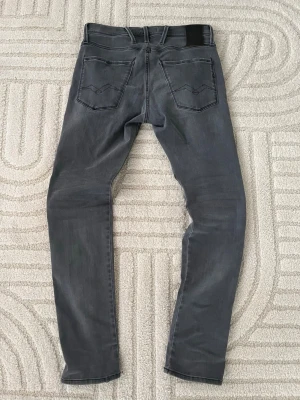 Grå  jeans från Replay - Säljer ett par grå jeans från Replay med slitningar  och klassiska bakfickor med broderad detalj. Jeansen har svarta sömmar och en svart patch bak i midjan. Materialet är stretchigt denim som sitter snyggt och bekvämt. Passar även w28-29