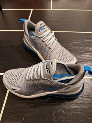 Nike Air Max 270 grå/blå sneakers - Nike Air Max 270 sneakers i grå mesh med blå detaljer och synlig Air-enhet i hälen. Skorna har en sportig siluett, ventilerande ovandel och klassisk Nike-logga på sidan och plösen. Perfekta för dig som gillar streetwear och bekväma sneakers.
