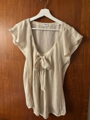 Krämvit vintage blus med rosett  - Söt och luftig krämvit vintage blus med korta, fladdriga ärmar och en dekorativ rosett framtill. Blusen har en rundad, djup urringning och är tillverkad i ett tunt, lätt tyg som ger en avslappnad känsla.