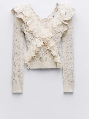 Krämvit stickad volangtröja Zara - Supersöt krämvit stickad tröja från Zara med volanger och spetsdetaljer längs v-ringningen och framtill. Tröjan har lång ärm, kortare passform och ett romantiskt mönster i garnet. Perfekt för dig som gillar vintage vibes och unika detaljer.