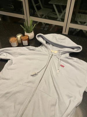 Ljusblå zip-hoodie från Levi's - Ljusblå hoodie med dragkedja från Levi's. Klassisk modell med huva och dragsko, två fickor framtill och liten röd Levi's-logga på bröstet. Perfekt för en chill och avslappnad stil. Mjuk bomullskänsla och enkel att matcha med jeans eller joggers.  Skriv för fler frågor 😃😃