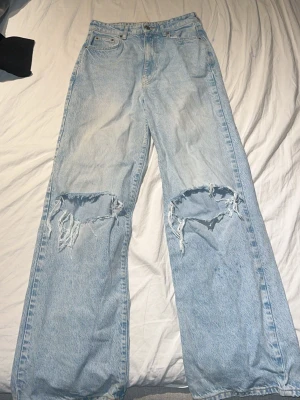 Ljusblåa baggy jeans med hål - Säljer ett par ljusblåa baggy jeans med stora slitna hål över knäna. Jeansen har hög midja, raka ben och klassisk femficksmodell. Perfekta för en avslappnad och trendig streetwear-look.