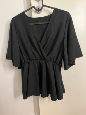 Svart omlottblus med vidd -  svart blus med v-ringning och markerad midja. Blusen har vida, korta ärmar. Ska ha ett band runt men är borta men tycker de funkar bra utan också.