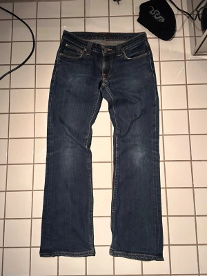 Bootcut jeans från Nudie Jeans - Säljer ett par klassiska mörkblå jeans från Nudie Jeans med bootcut längst ner och snygga kontrastsömmar. Jeansen har fem fickor och detaljerade broderier på bakfickorna. Tillverkade i slitstark denim, perfekta för en avslappnad och stilren look. Bootcut Eddy W28/L30 För små för mig men skulle nog sitta bra på dig som är mellan 170-175