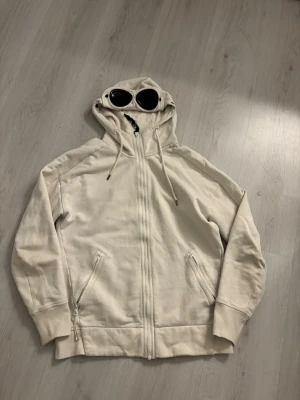 Beige C.P. Company hoodie med goggles - Säljer en beige hoodie från C.P. Company med dragkedja framtill och två fickor med dragkedja. Huvan har de ikoniska inbyggda goggles och justerbara snören. Perfekt för dig som gillar streetwear och unika detaljer.