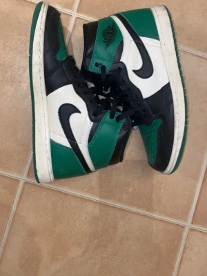 Nike Air Jordan 1 High svart/grön/vit - Säljer ett par Nike Air Jordan 1 High sneakers i svart, grönt och vitt läder. Klassisk siluett med svart swoosh, gröna detaljer och högt skaft. Perfekta för dig som gillar streetwear och vill sticka ut med en ikonisk modell.