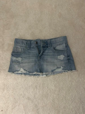 Ljusblå jeanskjol med slitningar - Kort jeanskjol i ljusblå denim med slitna detaljer och fransig nederkant. Kjolen har klassiska fickor fram och bak, bälteshällor och knappstängning. Perfekt för en avslappnad och trendig look.