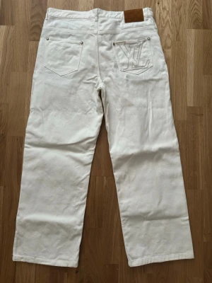 Louis Vuitton jeans - Säljer ett par exklusiva vita jeans med raka ben och klassisk passform från Louis Vuitton i storlek 31. Mycket fint skick och har endast använts ett par gånger då den är för kort för mig. Byxorna har klassiska LV-detaljer med guldiga knappar och nitar, samt det ikoniska blå/vita Damier-mönstret på insidan av fickorna och linningen. Bakfickan har en stor LV-logga i beige brodyr och ett brunt läderpatch i midjan. Perfekta för dig som vill sticka ut med exklusiva detaljer och stilren design.