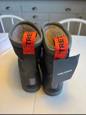 Svarta fodrade stövlar från Tretorn - Säljer ett par svarta stövlar från Tretorn med varm, grått foder och tydliga röda detaljer på hälen. Stövlarna har en robust design och är perfekta för kalla och blöta dagar. Tillverkade i slitstarkt gummimaterial och har en bekväm passform. Nya med prislapp. Storlek: 32, Innermått: 21,5cm