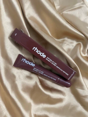 rhode Peptide Lip Tint PBJ - Säljer ett rhode Peptide Lip Tint i färgen PBJ. Läppbalsamet kommer i en mörklila tub med matchande kartong och har en mjuk applikator. Produkten är berikad med peptider och ger en lätt tonad finish. Perfekt för dig som vill ha återfuktade och snygga läppar.