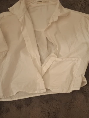 Vit kortärmad skjorta från Pull&Bear - Stilren vit skjorta från Pull&Bear i bomull. Skjortan har korta ärmar, klassisk krage och knappar framtill. Perfekt för en avslappnad och trendig look. Enkel att matcha med olika outfits.
