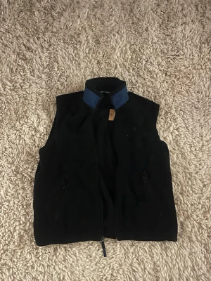 Svart fleeceväst från Ralph Lauren - Snygg svart fleeceväst från Ralph Lauren med blått broderat logotyp på bröstet och kontrasterande blå krage. Västen har dragkedja framtill och två fickor med dragkedja. Perfekt att slänga över en hoodie eller tröja för extra värme.