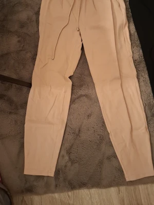 Beige och svarta mjukisbyxor - Två par mjukisbyxor, ett par i beige och ett par i svart. Båda har dragsko i midjan med metalländar och smal passform. Perfekta för chill dagar eller när du vill vara bekväm. Materialet ser ut att vara mjukt och stretchigt, troligen bomull eller bomullsblandning.