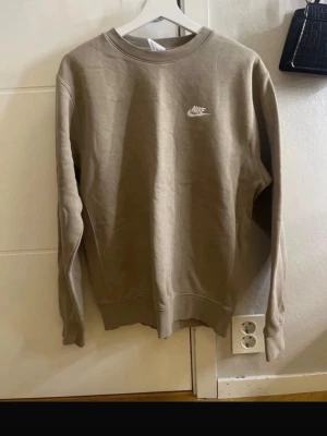 Beige sweatshirt från Nike - Säljer en beige sweatshirt från Nike med klassisk rund halsringning och vit Nike-logga på bröstet. Tröjan har långa ärmar och är tillverkad i mjukt material med borstad insida för extra komfort. Perfekt för en avslappnad och sportig stil.