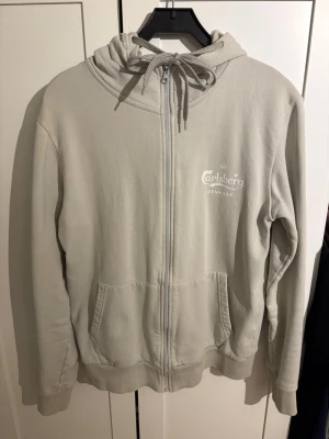 Carlsberg zip hoodie - Beige hoodie med dragkedja från Carlsberg. Har huva med dragsko, två fickor framtill och tryckt logga på bröstet. Perfekt för en chill och avslappnad stil. Text på armen är lite sliten annars i bra skick.