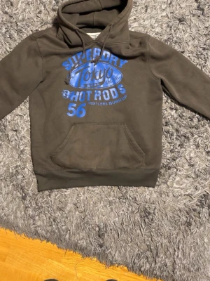 Mörkgrön Superdry hoodie – storlek L - Mörkgrön hoodie från Superdry med blått tryck framtill. Storlek L. Skön kvalitet och bra skick.