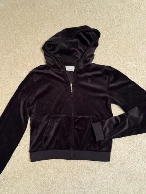 Svart velourhoodie från Juicy Couture - Mjuk och lyxig svart velourhoodie från Juicy Couture med J-hänge som dragkedja framtill, huva och broderad JC-logga på bröstet. Två fickor framtill och ribbade muddar vid ärmslut och nederkant. I mycket fint skick, sparsamt använd. Nypris 950kr