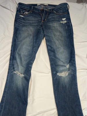 Blå slitna jeans från Abercrombie & fitch - Säljer ett par blå jeans från Abercrombie and fitch med snygga slitningar på knäna och låret. Jeansen har klassisk femficksmodell, låg midja och utsvängda ben. Perfekta för en avslappnad och trendig look.