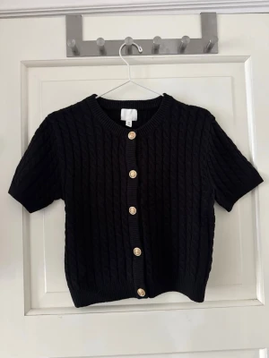 Kabelstickad topp - Svart kabelstickad topp med guldknappar. Från H&M, storlek M. Ny utan prislapp, aldrig använd. 60% bomull och 40% akryl. Skickar med en extra knapp. Lite kortare modell. 