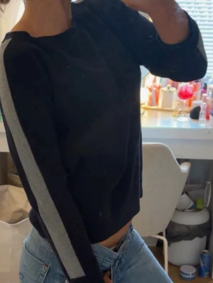 Mörkblå  tröja med grå ränder på ärmarna - Snygg svart tröja med raka grå ränder längs båda ärmarna. Modellen är rak och har rund halsringning. Perfekt till jeans för en avslappnad och trendig look.