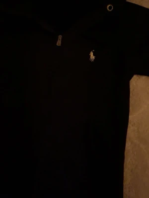 Svart hoodie från Ralph Lauren - Svart hoodie med dragkedja vid halsen och klassisk broderad logga på bröstet. Mjuk och skön med huva och långa ärmar, perfekt för en chill och stilren look.