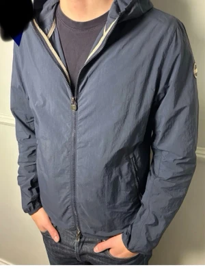 Blå vindjacka från Colmar - Snygg blå vindjacka från . Colmar med dragkedja framtill, huva och två sidofickor med dragkedja. Jackan har en diskret logotyp på ärmen och är tillverkad i lätt, vindtätt material som passar perfekt för blåsiga dagar. Passar 180-182 8/10 passform.