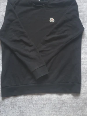 Svart stickad tröja från Moncler - Svart stickad tröja från Moncler med rund halsringning och Moncler-logga broderad på bröstet. Tröjan har långa ärmar och ribbade muddar vid ärmslut och nederkant. Perfekt för en stilren och enkel look. NFC funkar. Äkta tröja som jag fick som present för c.a 4,000kr.