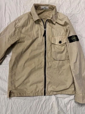Beige overshirt från Stone Island - Stilren beige overshirt från Stone Island med klassisk svart och gul logotyp på ärmen. Jackan har en rak passform, dragkedja framtill och en bröstficka med knapp. Perfekt lager på lager-plagg för en trendig streetwear-look.