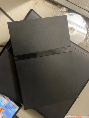 Sony PlayStation 2 Slim svart konsol - Säljer en svart PlayStation 2 Slim från Sony. Konsolen har en tunn och stilren design med mattsvart finish och en blank remsa där PS2-loggan sitter. Perfekt för dig som vill spela klassiska PS2-spel och njuta av retro gaming.