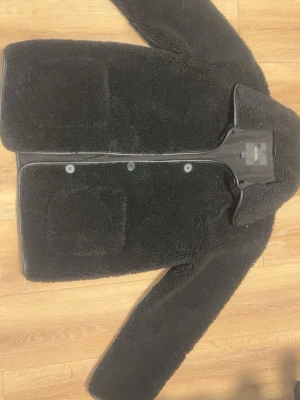 Svart teddyjacka från Monki - Mysig svart teddyjacka från Monki med stora framfickor och knäppning framtill. Jackan har klassisk krage och är tillverkad i ett fluffigt teddy-material som håller dig varm. Perfekt för kyliga dagar och enkel att styla till olika outfits.