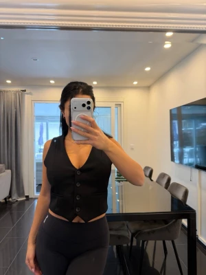 Svart figursydd väst med knappar - Stilren svart väst med djup V-ringning och tre knappar framtill. Figursydd modell med markerad midja och klassisk krage. Perfekt att styla över en t-shirt eller skjorta för en trendig look.