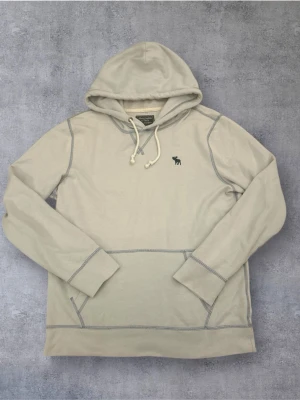 Abercrombie & Fitch Hoddie - Snygg beige hoodie med huva och dragsko från Abercrombie & Fitch. Har en liten mörk logga broderad på bröstet och kontrastsömmar. Tillverkad i mjukt bomullsmaterial och har en klassisk känguruficka framtill.