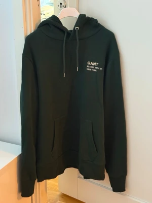 Svart hoodie från GANT med tryck - Snygg svart hoodie från GANT med vitt tryck på bröstet där det står 'GANT 52 EAST 39TH ST. NEW YORK'. Hoodien har dragsko i huvan och en stor magficka. Perfekt för en chill och stilren look.