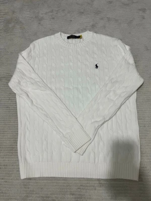 Vit kabelstickad tröja Polo Ralph Lauren - Kabelstickad vit tröja från Polo Ralph Lauren med rund halsringning och den klassiska marinblå logotypen broderad på bröstet. Tröjan har ribbade muddar vid ärmslut och nederkant. Perfekt för en clean och stilren look.