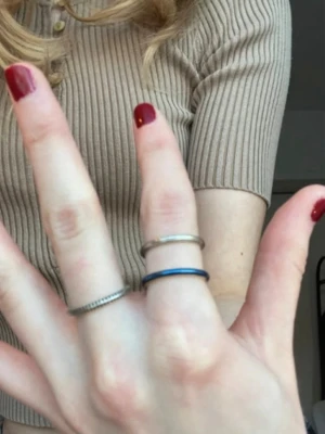 Set med ringar från Pandora - Säljer ett set med tre superfina ringar från Pandora i minimalistisk design. De alla består av sterling silver. En har små svarta kristaller runt sig men ser mest silvrig ut, den andra har små vågräta linjer/ränder i sig men silverfärgad den med. Den sista har ett tjockare blått streck ovanpå silvret. Perfekta att bära tillsammans eller var för sig för en trendig look.