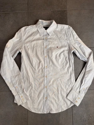 Vitblå randig skjorta från Gant - Klassisk vit och ljusblå randig skjorta från Gant med broderad logga på bröstet. Skjortan har lång ärm, knappar framtill och är tillverkad i bomull. Perfekt för en stilren och avslappnad look.