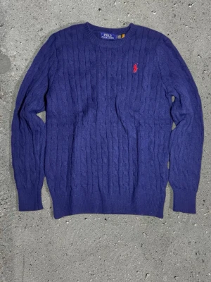 Blå kabelstickad tröja Polo Ralph Lauren - Kabelstickad blå tröja från Polo Ralph Lauren med rund halsringning och röd broderad logga på bröstet. Tillverkad i 100% bomull och har ribbade muddar vid ärmslut och nederkant. Perfekt för en stilren och klassisk look.