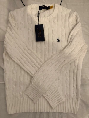 Vit kabelstickad tröja Polo Ralph Lauren - Vit kabelstickad tröja från Polo Ralph Lauren med rund halsringning och broderad logga på bröstet. Tröjan har ribbade muddar vid ärmslut och nederkant. Klassisk och stilren design som passar till mycket.