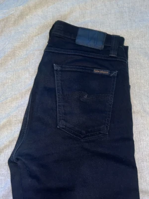 Mörkblå jeans (Nudie) - Snygga svarta jeans från Nudie Jeans med klassisk femficksmodell och diskret logga på bakfickan. Jeansen har normal passform och är tillverkade i mjuk bomull med en stilren look. Perfekta för dig som gillar enkel och clean stil. Aldrig använda helt nya!