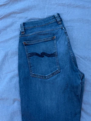 Blå jeans från (Nudie) - Säljer ett par klassiska blå jeans från Nudie Jeans med snyggt vågmönster på bakfickan. Jeansen har fem fickor, normal midja och raka ben. Tillverkade i mjuk denim som ger en avslappnad look. Perfekta för dig som gillar stilrena och tidlösa jeans. Bra skick W. 31 L. 32