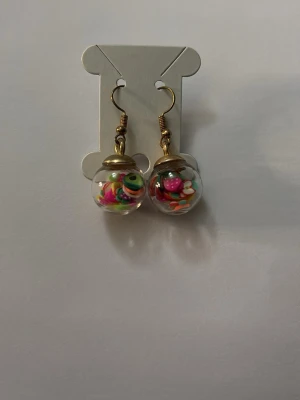 Glaskulor örhängen med färgglada pärlor - Unika örhängen med glaskulor fyllda med små, färgglada pärlor i olika former och nyanser. Krokarna är i guldfärgad metall och designen är lekfull och kreativ – perfekt för dig som gillar statement-accessoarer.